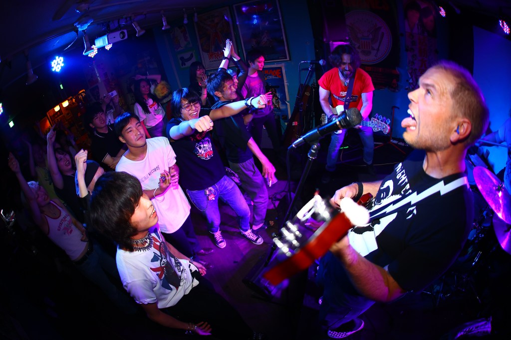 A lively music performance in a dimly lit venue with colorful lights, featuring a band playing on stage and an enthusiastic crowd dancing and cheering.
