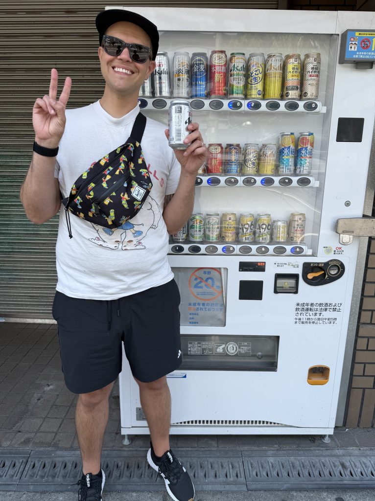 A person smiling and making a peace sign while holding a can of drink in front of a vending machine filled with various canned beverages.