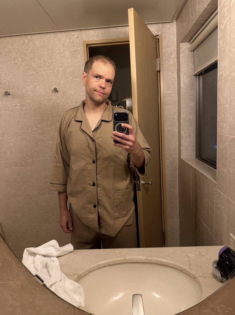 A person wearing a beige pajama set stands in front of a bathroom mirror, holding a smartphone for a selfie. The bathroom features a sink, a towel, and a door partially open in the background.