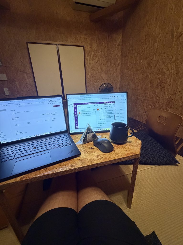 A workspace setup featuring two laptops on a wooden table, a coffee mug, a mouse, and a triangular snack, with a person seated cross-legged in shorts. The room has wooden walls and a fan in the background.