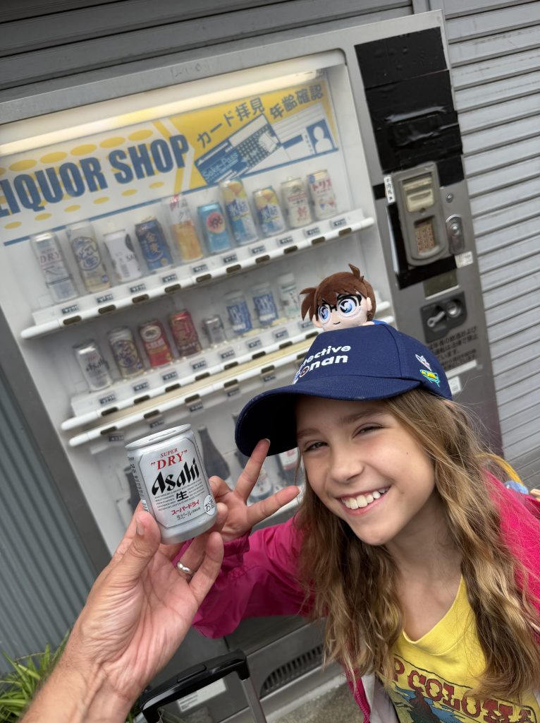 A young girl smiling while holding a can of Asahi beer in front of a vending machine displaying various beverages in a 'Liquor Shop' setting.