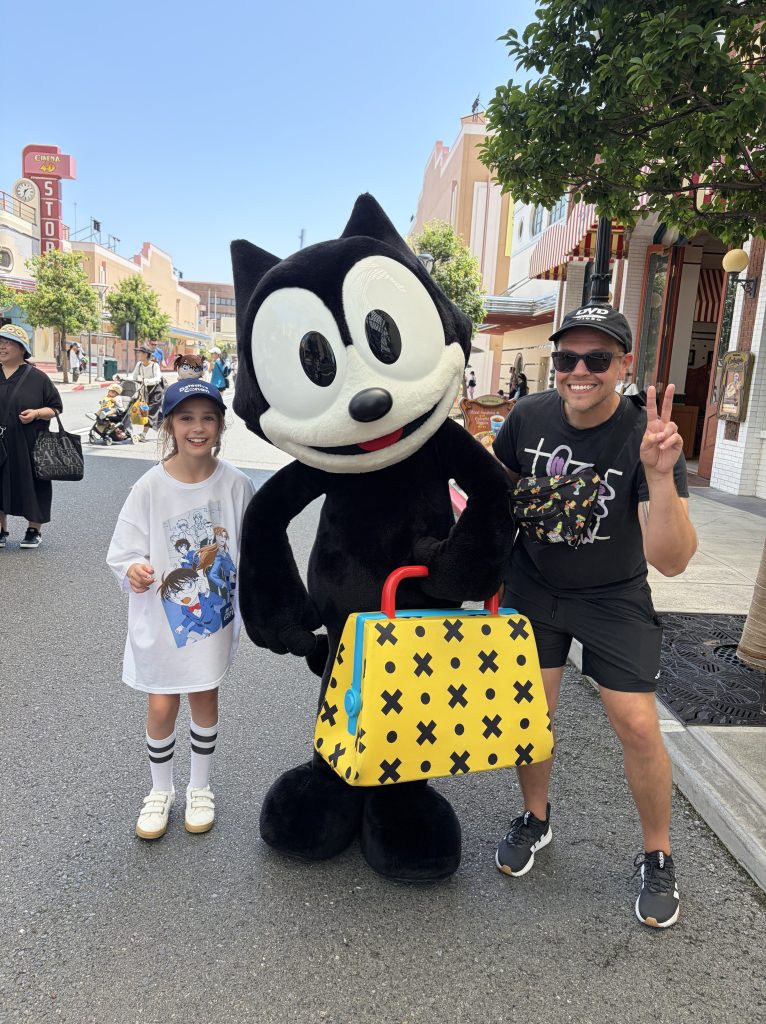 A young girl wearing a cap and a t-shirt poses with a large cartoon character in a black outfit, holding a colorful handbag. A man wearing sunglasses and a black shirt is making a peace sign next to them in a bustling outdoor setting.