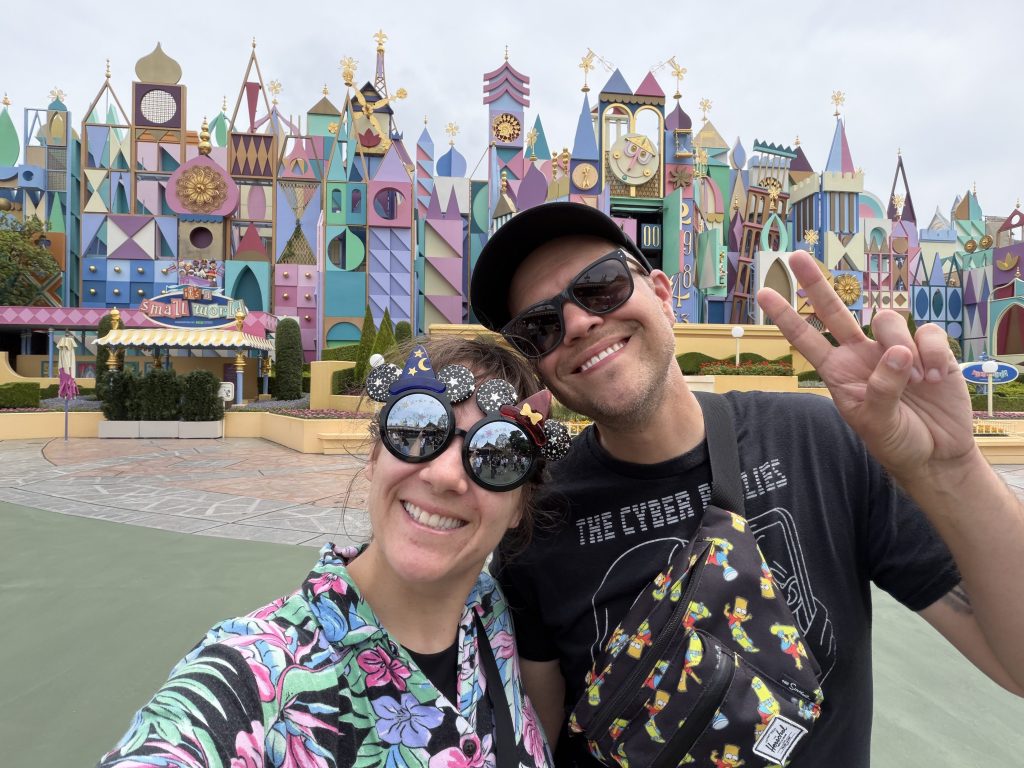A smiling couple stands in front of the colorful 'It's a Small World' attraction at a theme park, with vibrant, whimsical architecture in the background.